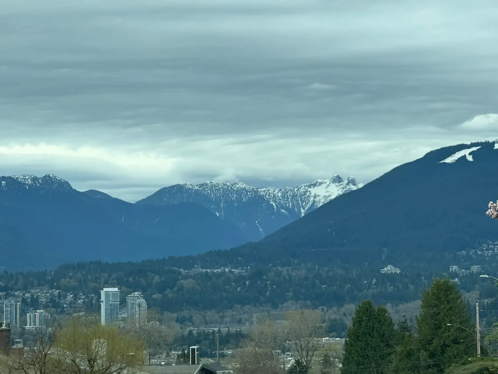 View of the Lions mountains with light snowpack overlooking North Vancouver residential areas and buildings.