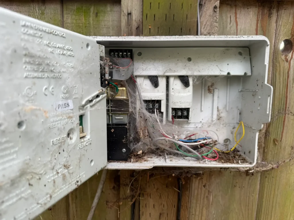 An old, neglected Rain Bird irrigation controller covered in cobwebs and dirt inside an outdoor box.