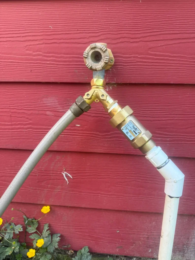 An outdoor hose bib faucet with a brass Y-splitter connecting a grey garden hose and an Apollo dual-check backflow preventer assembly leading into a white PVC drip irrigation pipe against a red wood wall.