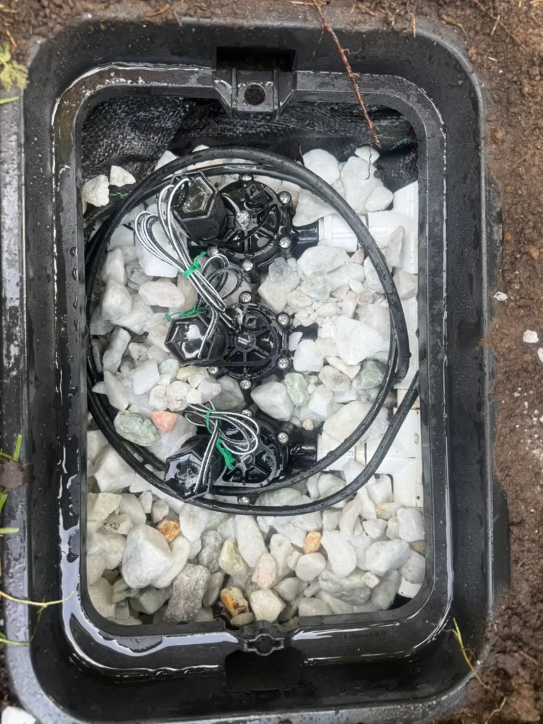 An overhead view of an open black plastic valve box installed in the ground, containing three automatic inline irrigation valves surrounded by white gravel or marble chips with coiled low-voltage wiring.